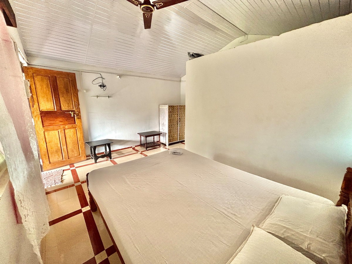 A serene sleeping area features a spacious bed with white bedding against a light-colored wall. Two small black tables are positioned on either side, with wooden accents visible in the room. Soft natural light enters through an opening, illuminating the inviting space.