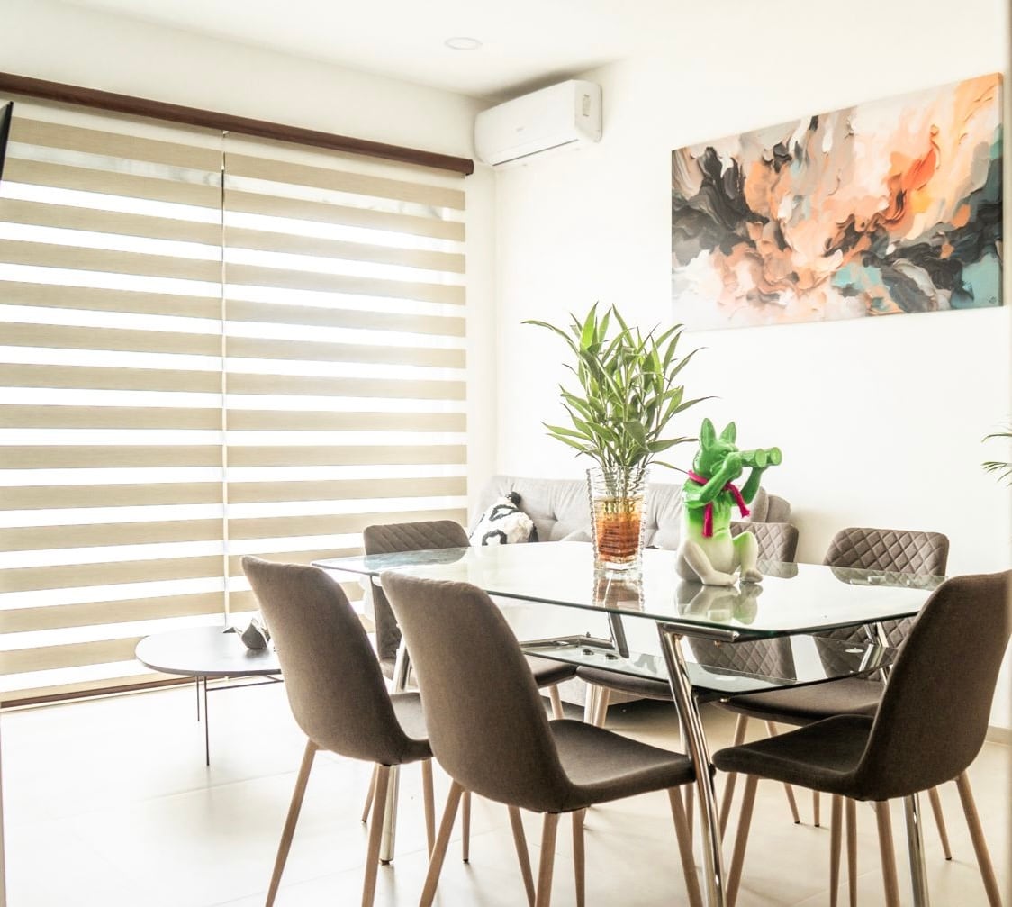 A bright dining area is presented, featuring a glass table surrounded by six upholstered chairs. A potted plant is placed at the center of the table, while a modern artwork adorns the wall above a comfortable sofa, complemented by large windows with layered shades.
