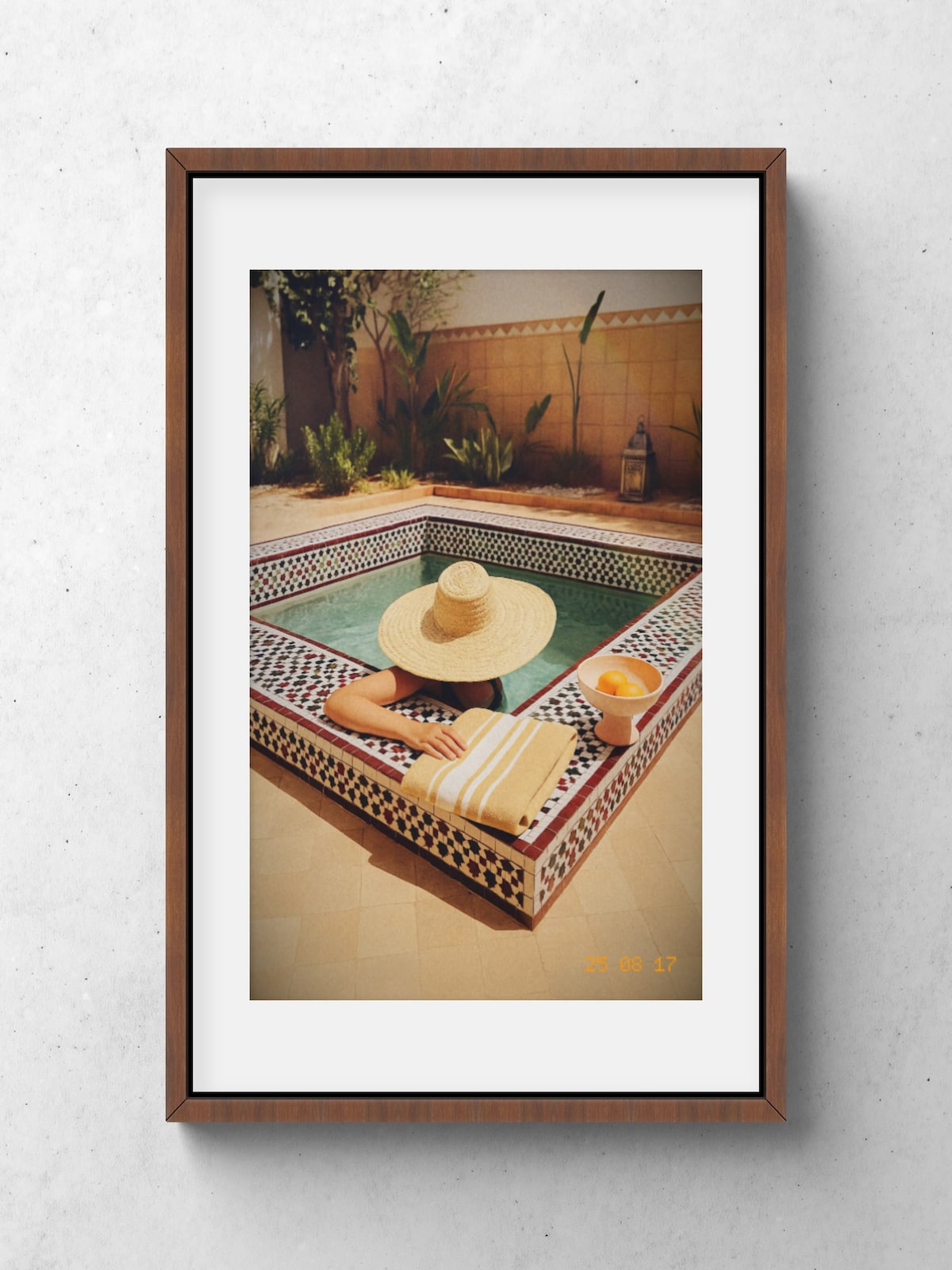 A serene pool area features a small tiled pool surrounded by decorative tiles and lush greenery. A person in a wide-brimmed hat lounges on the edge, with a striped towel draped beside them and a bowl of fruit placed nearby.