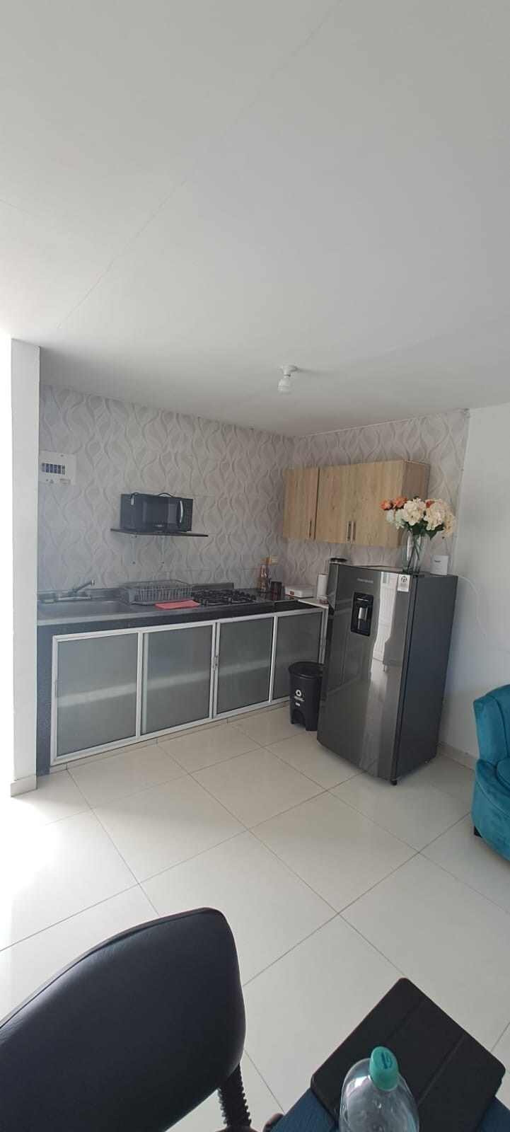 A modern kitchen area is visible, featuring a sleek gas stove and a double-door refrigerator. Upper cabinets provide storage, with a light-colored backsplash complementing the wooden cabinetry. A turquoise armchair adds a splash of color to the neutral-toned space, enhancing its functionality.