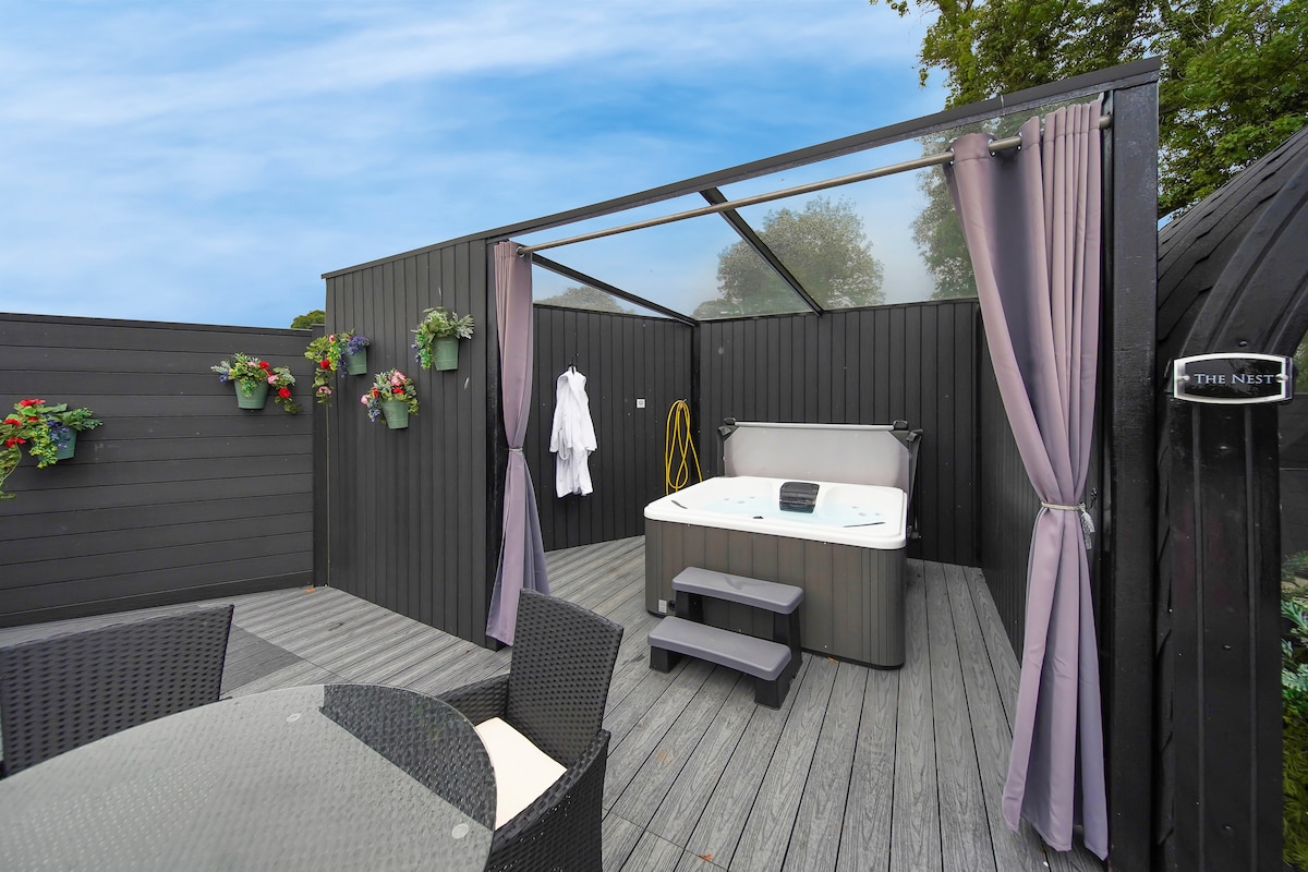 An outdoor area is depicted, featuring a hot tub surrounded by wooden decking. Potted plants are lined along the walls, adding greenery. A table with chairs is visible, with light curtains framing the space. A bathrobe hangs nearby, hinting at relaxation options.