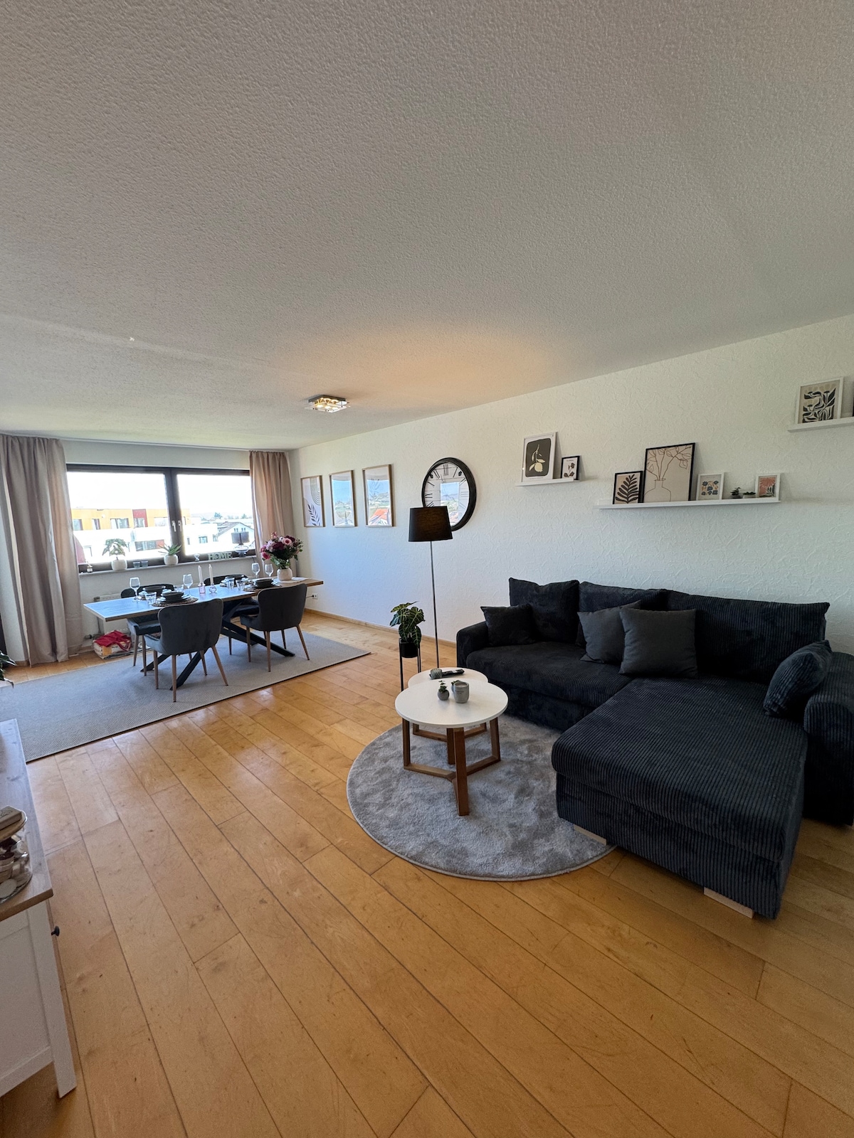 A bright living area features a plush dark sofa with decorative pillows, arranged on a circular area rug. A round coffee table sits in front, complemented by a floor lamp. Nearby, a dining table with chairs is set against a large window, allowing natural light to flow in.