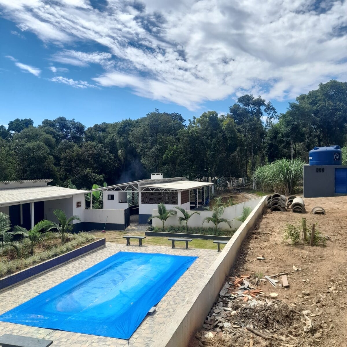 A spacious outdoor area is highlighted by a blue pool covered with a protective tarp. Surrounding greenery can be seen in the background, along with well-maintained buildings that offer leisure facilities. The setting combines natural elements with functional spaces for relaxation and recreation.