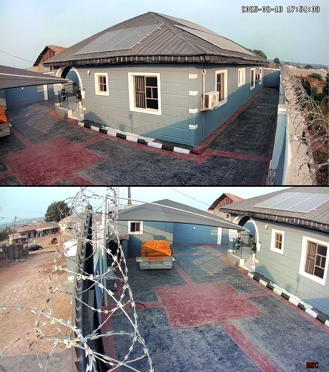 A well-maintained exterior of a single-story building is displayed, featuring light blue siding and a protective wrought iron fence. The paved area is spacious, with a combination of red and gray pathways, and a large orange couch is visible under an awning.