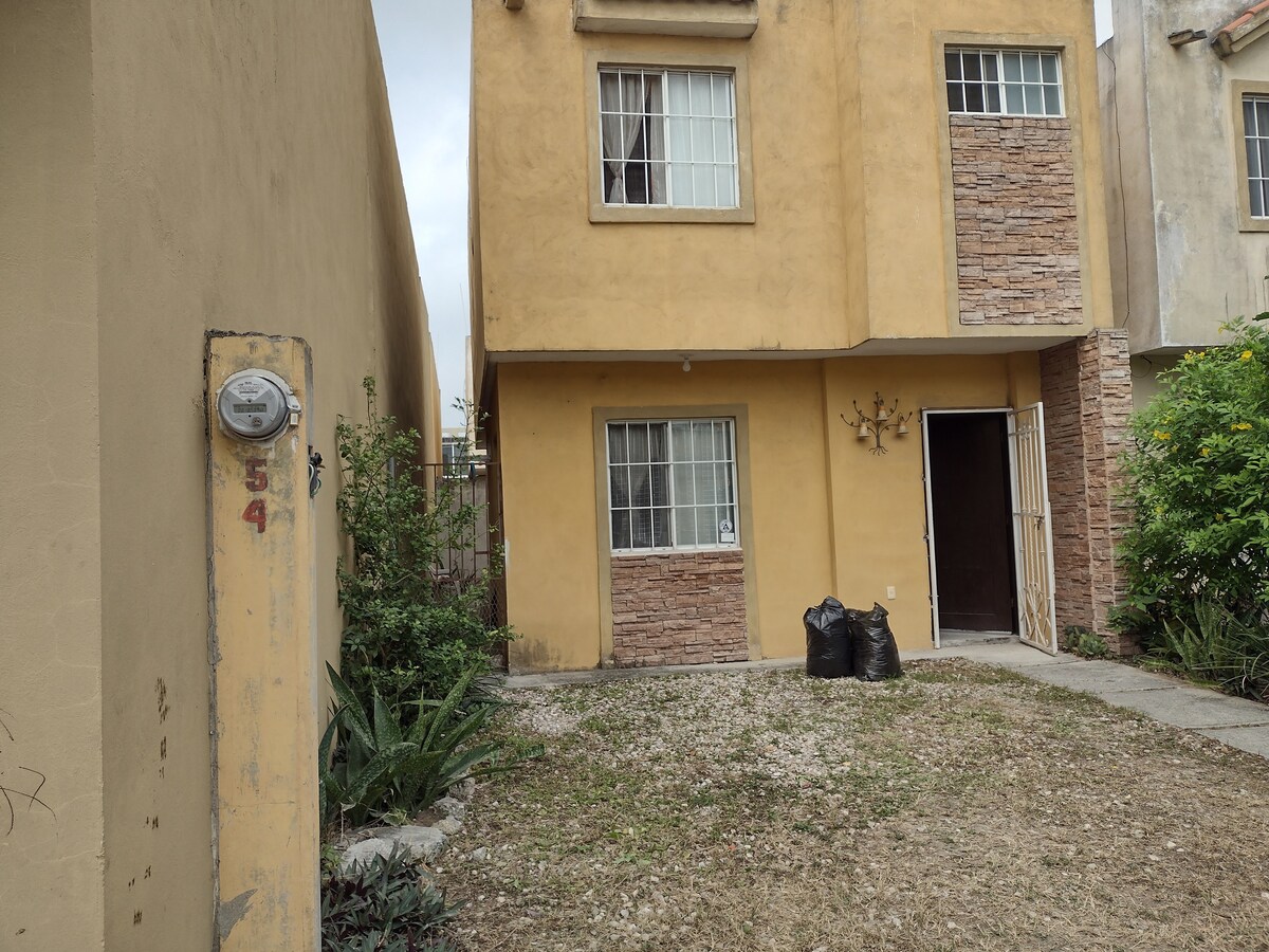 A two-story building features a textured yellow facade complemented by stone accents. The front entrance is framed by a large window, and the area is bordered by gravel and greenery. Two black bags are positioned near the doorway, suggesting an outdoor space that is easily accessible.