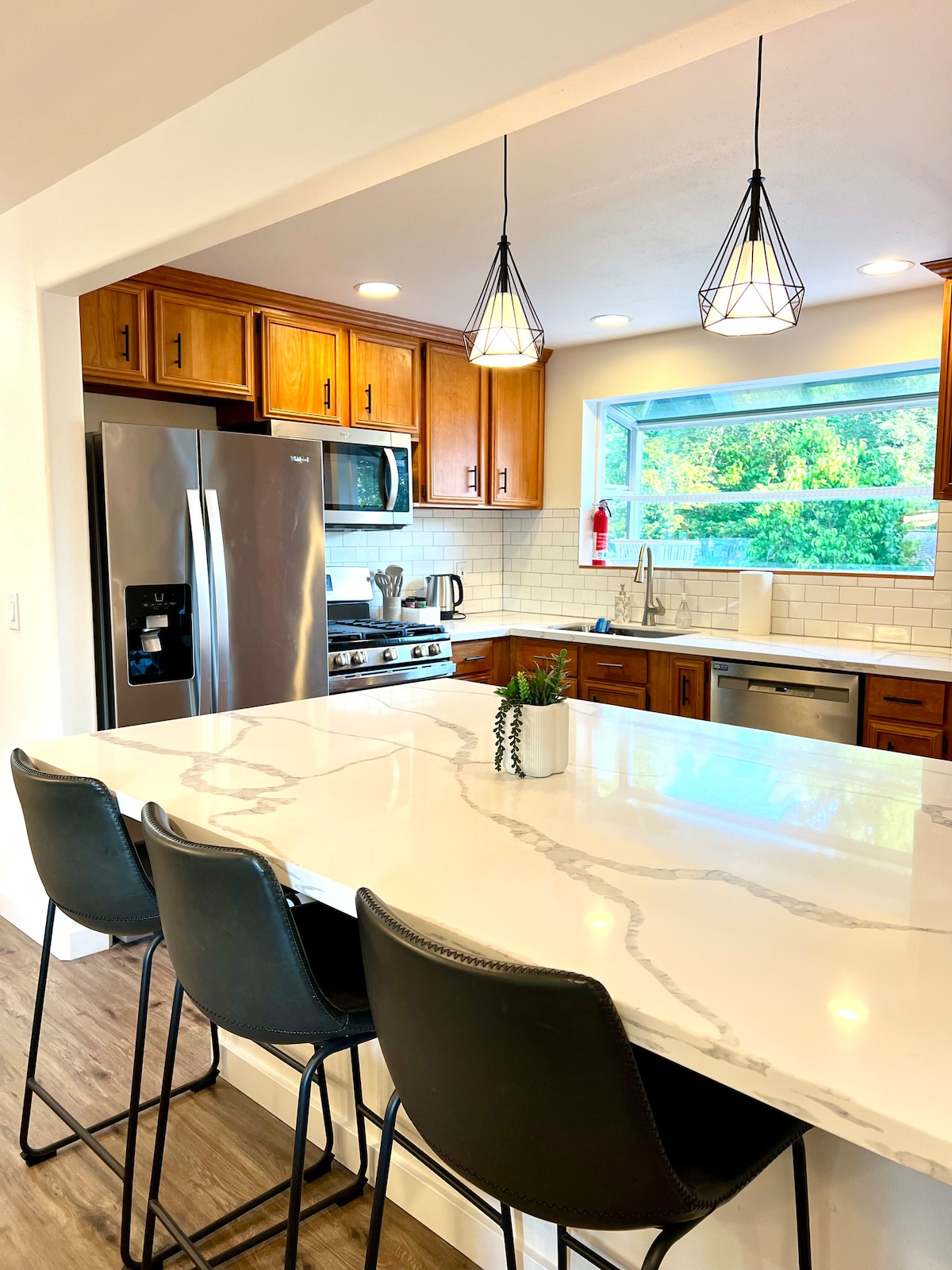 The kitchen features modern cabinetry, stainless steel appliances, and a spacious countertop with seating for three. Natural light enters through a window, highlighting the clean lines and neutral tones of the decor. Decorative pendant lights hang above the counter.