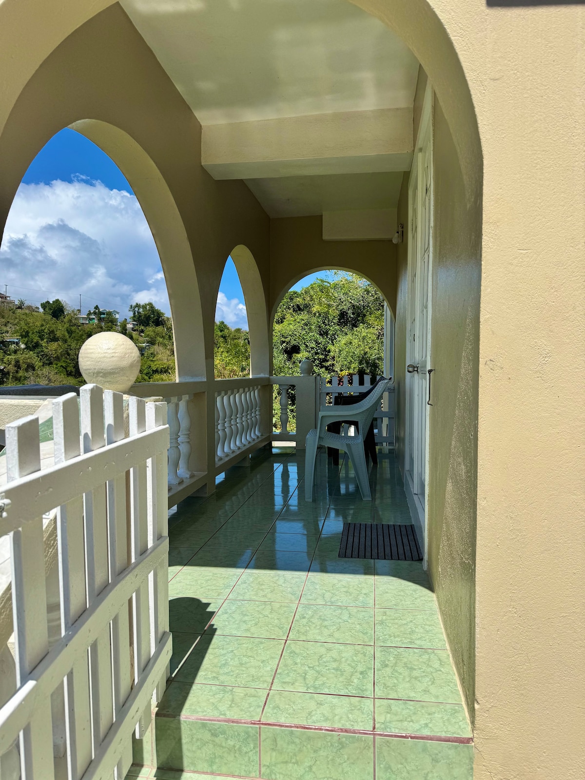A private balcony is showcased with a view through arched openings. The space is adorned with pale green tiles and features a white chair positioned beside a small table. Bright sunlight enhances the serene atmosphere, while lush greenery can be seen in the background.