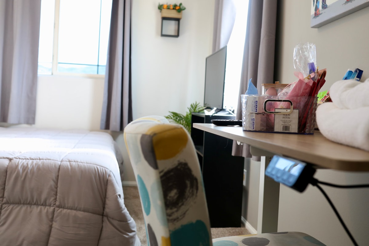 A cozy private room is presented, featuring a plush California King bed with soft linens. A desk with a colorful chair is set beside the bed, holding complimentary treats. Natural light fills the space through a nearby window, with blackout curtains partially drawn.