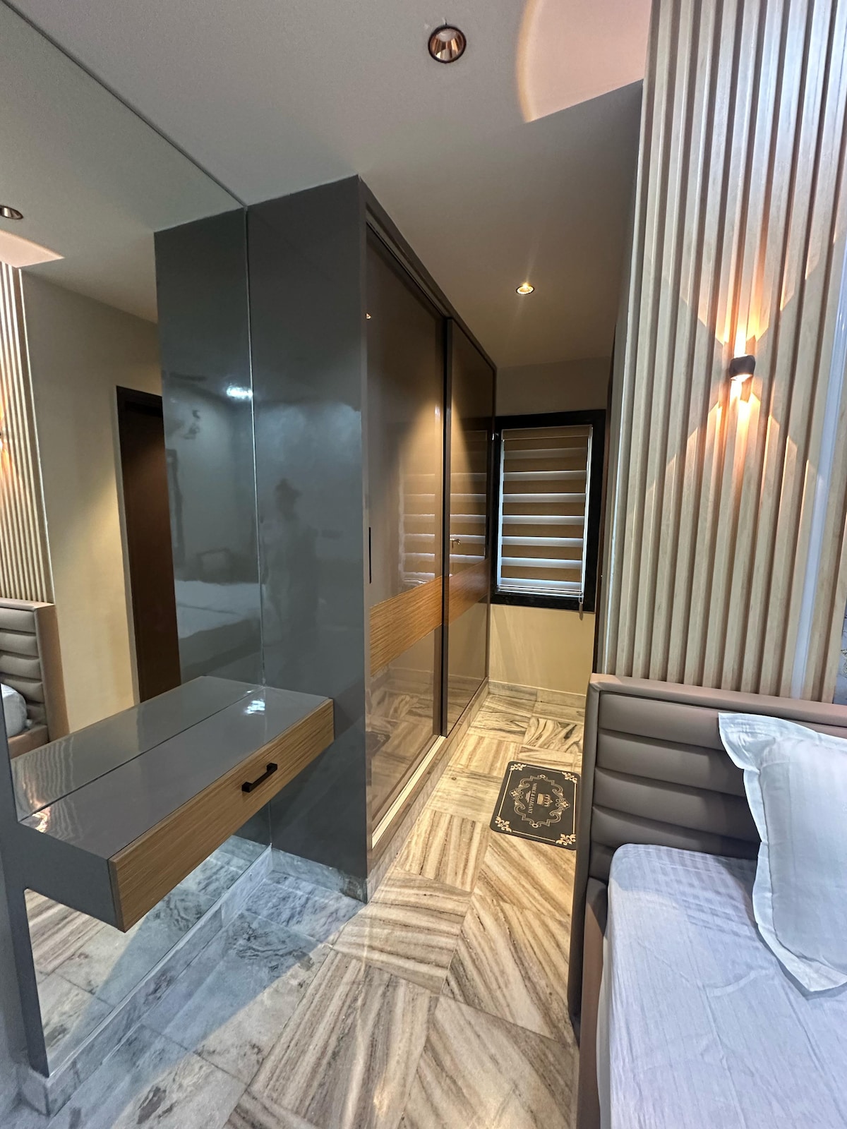 A modern interior features sleek, mirrored wardrobes and elegant wooden accents. Soft lighting highlights the walls, while a small console table offers a practical touch. The flooring showcases a stylish tile pattern, contributing to the room's overall contemporary design.