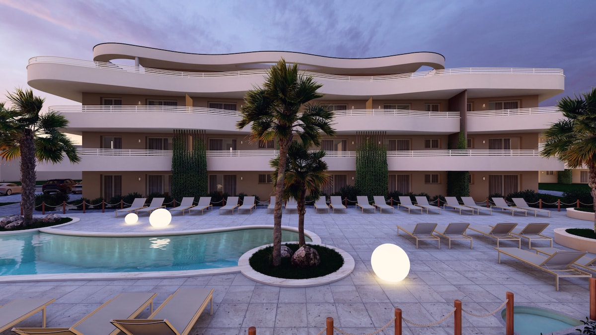 A modern building with multiple levels is surrounded by palm trees and lounge chairs. A curvy balcony above enhances the architectural design. A circular pool with illuminated spheres is visible in the foreground, creating a welcoming outdoor space for relaxation.