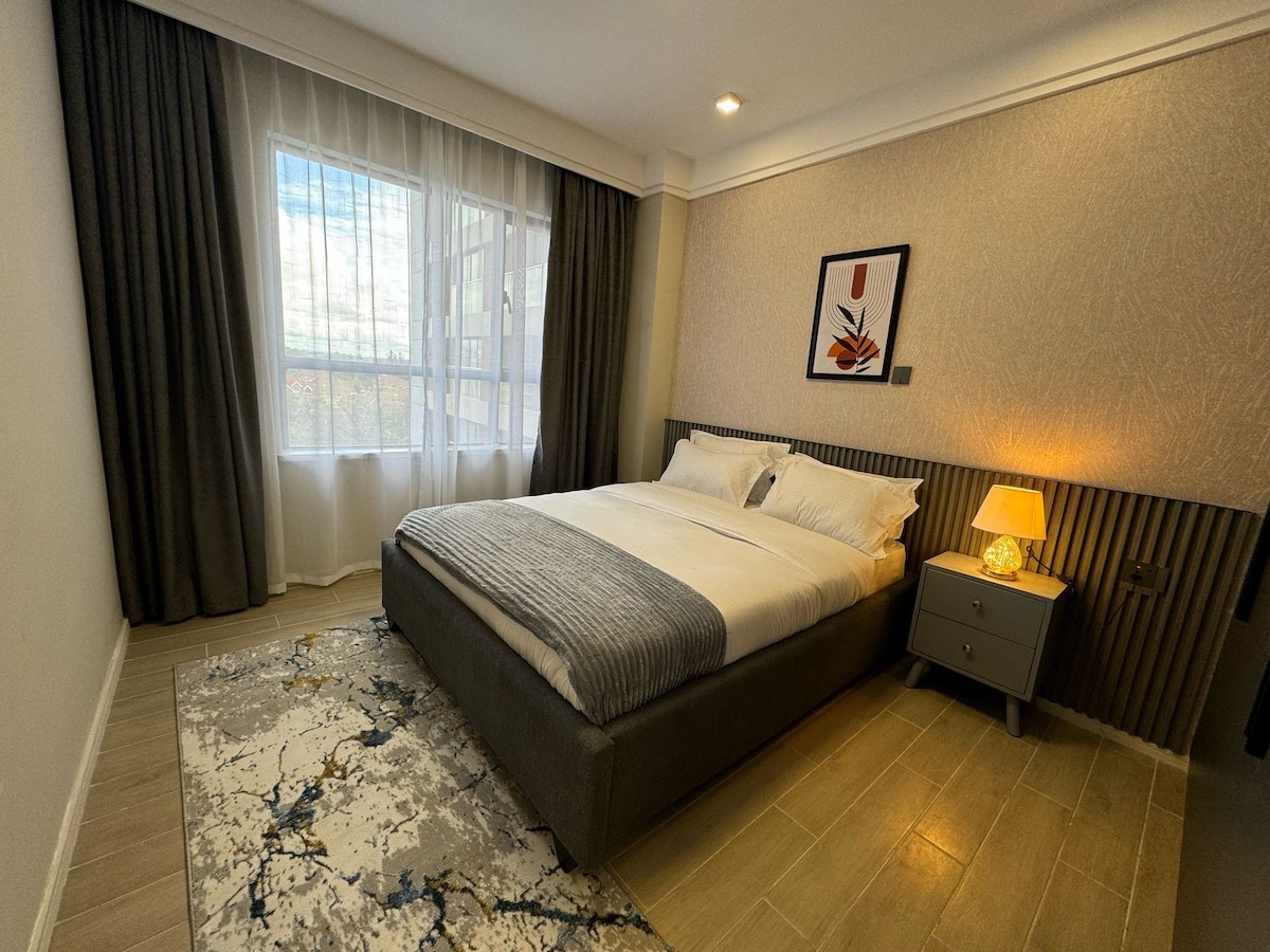 A cozy sleeping area is presented with a spacious bed dressed in crisp white linens. A bedside table with a lamp is positioned next to the bed. Natural light filters through a large window, covered by sheer curtains, illuminating the neutral-toned walls and patterned rug.