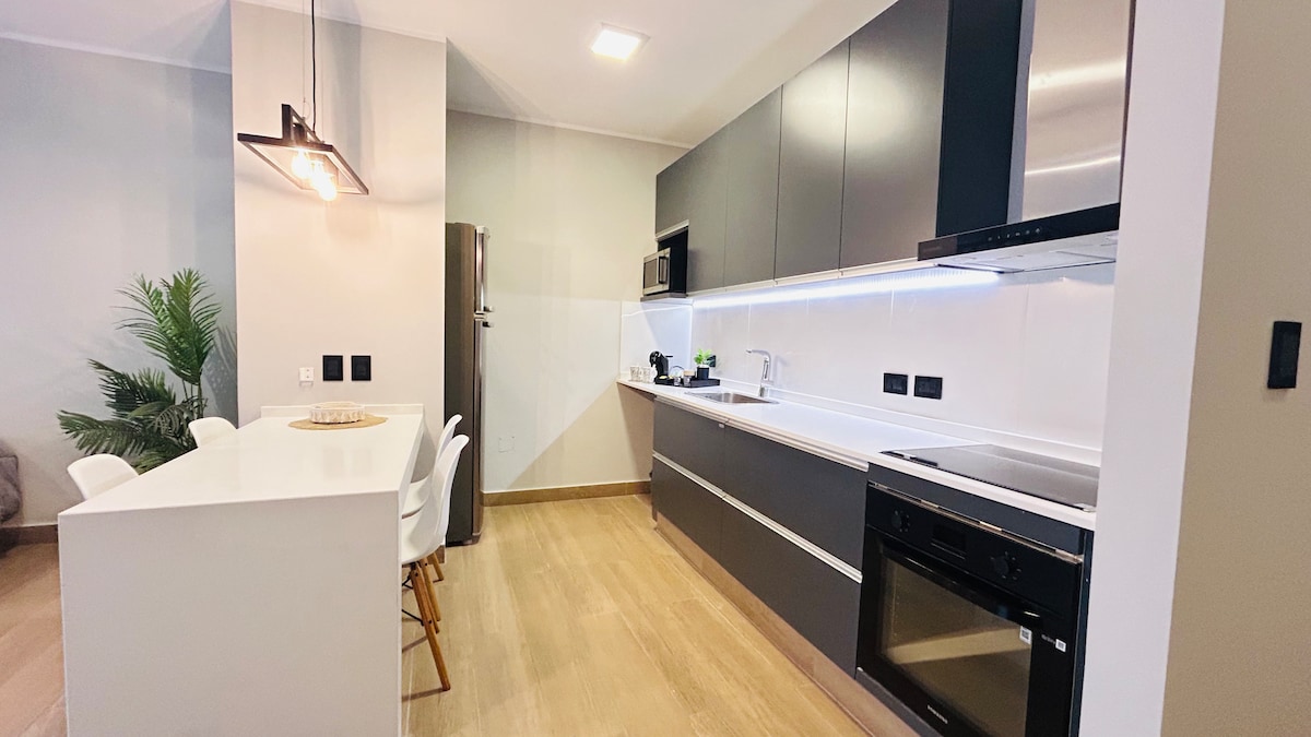 A modern kitchen is displayed with sleek cabinetry and a white countertop. Essential appliances, including a stove and refrigerator, are visible. A dining area with light-colored chairs is nearby, complemented by a decorative plant in the corner, enhancing the room's functionality and style.