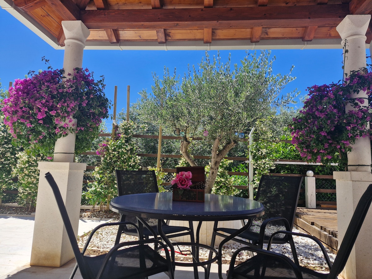 A private outdoor terrace is surrounded by vibrant flowers hanging from stone pillars. A round table with black chairs invites relaxation. Lush greenery, including an olive tree, provides a serene backdrop, enhancing the peaceful atmosphere of the space.