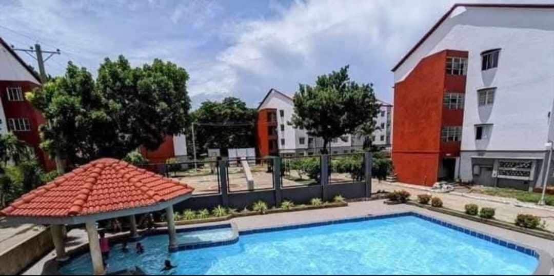 A clear blue pool is surrounded by a tiled area with shaded seating. Lush trees provide greenery in the background, while contemporary buildings with red accents stand in proximity. An outdoor area designed for recreation is visible, enhancing the inviting atmosphere.