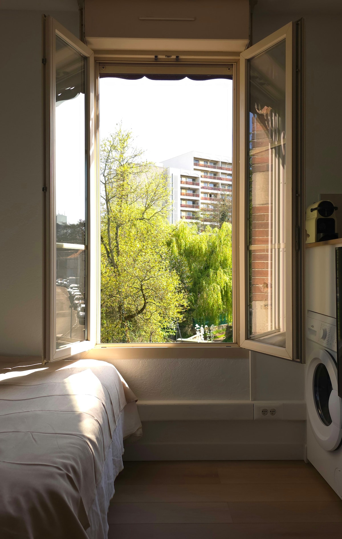 A window is opened wide, allowing natural light to fill the room. Outside, lush trees provide greenery, while a modern building is visible in the background. The soft light complements a neatly made bed situated beside the window.