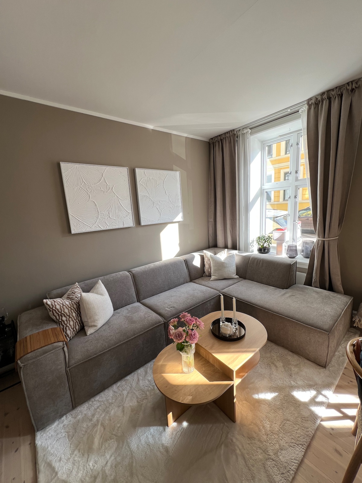 A spacious living area features a light gray sectional sofa with decorative pillows. A round coffee table is centered in front, adorned with a candle and a small vase of flowers. Natural light filters through a large window, complemented by soft curtains, creating a calming space.