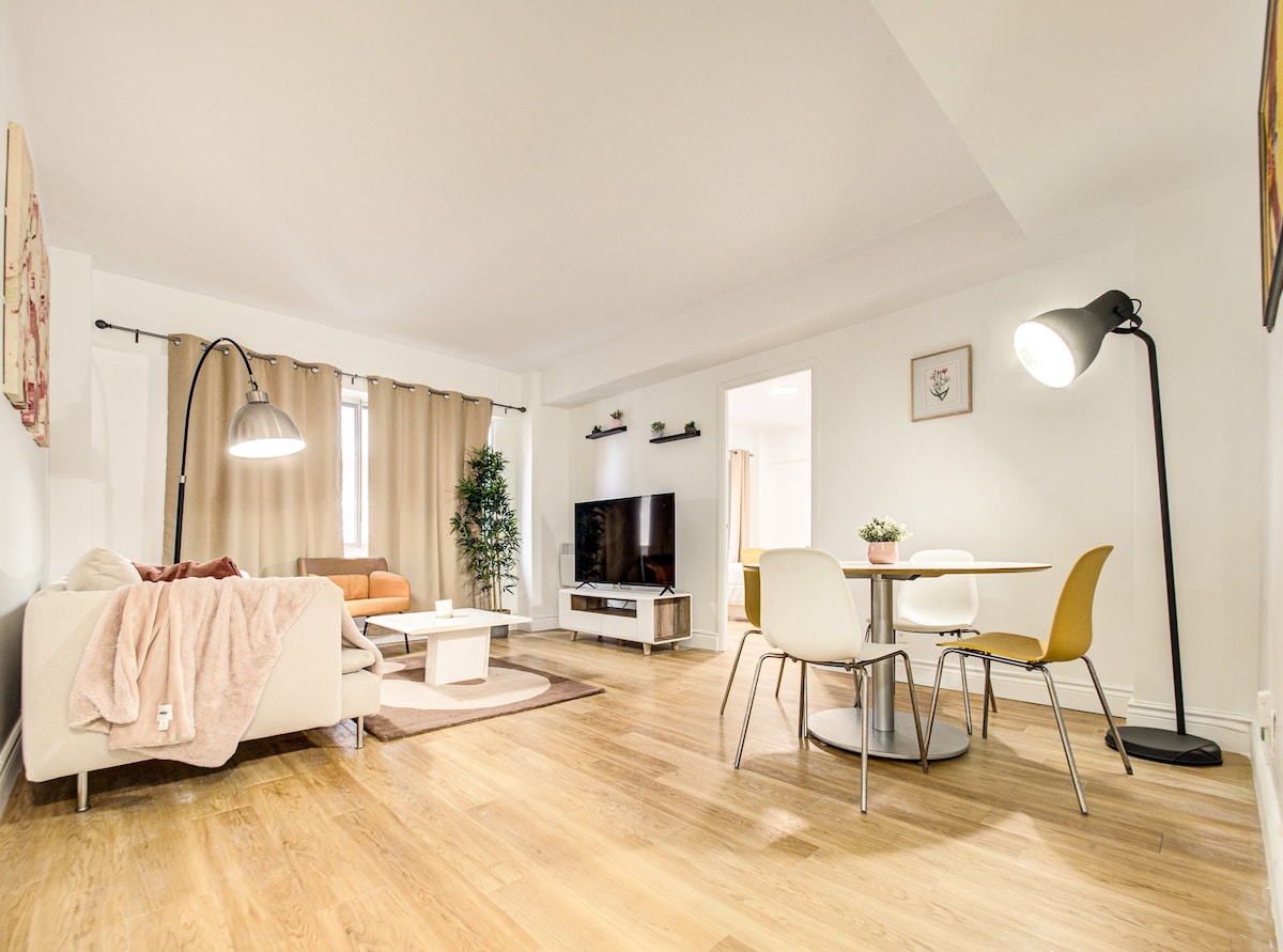 A contemporary living space showcases a blend of comfortable seating, including a bright orange couch and a beige armchair. A wooden dining table surrounded by modern chairs is positioned near a floor lamp. A large TV is mounted on the wall, complemented by warm lighting from nearby fixtures.