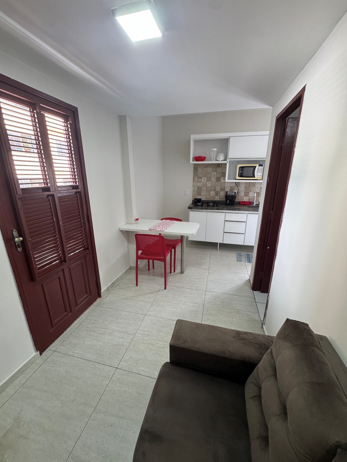 A compact kitchen area is visible, featuring a small dining table with red chairs and essential appliances. Light-colored tiles and walls create a bright atmosphere. A brown sofa is positioned in the corner, providing a comfortable seating option. Traditional wooden doors with slatted design add character.
