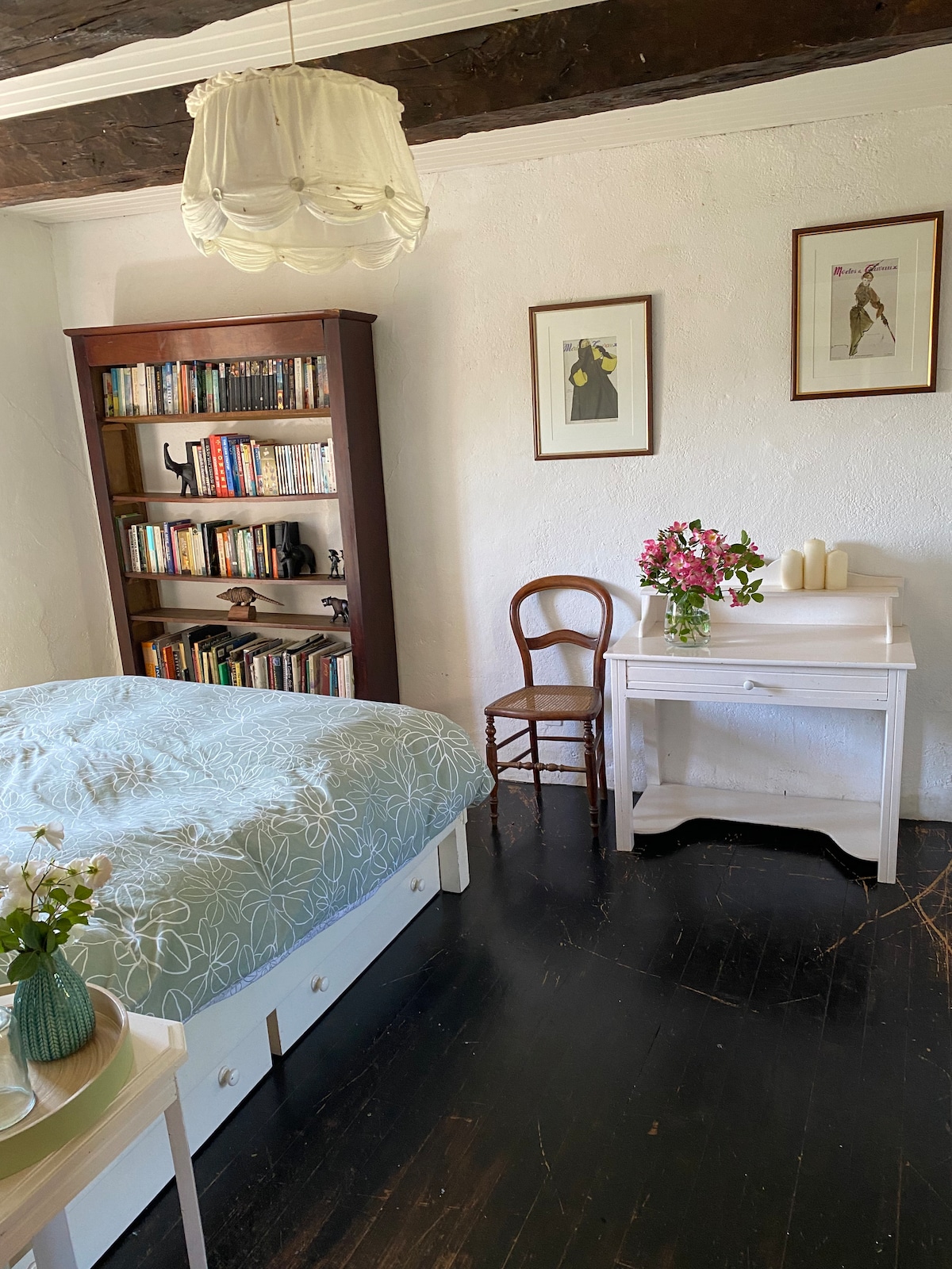 A serene bedroom features a spacious bed with a light blue coverlet, complemented by a wooden bookshelf filled with books. A small, white writing desk sits beside a wooden chair, adorned with a bouquet of flowers and candles, creating a cozy reading nook.