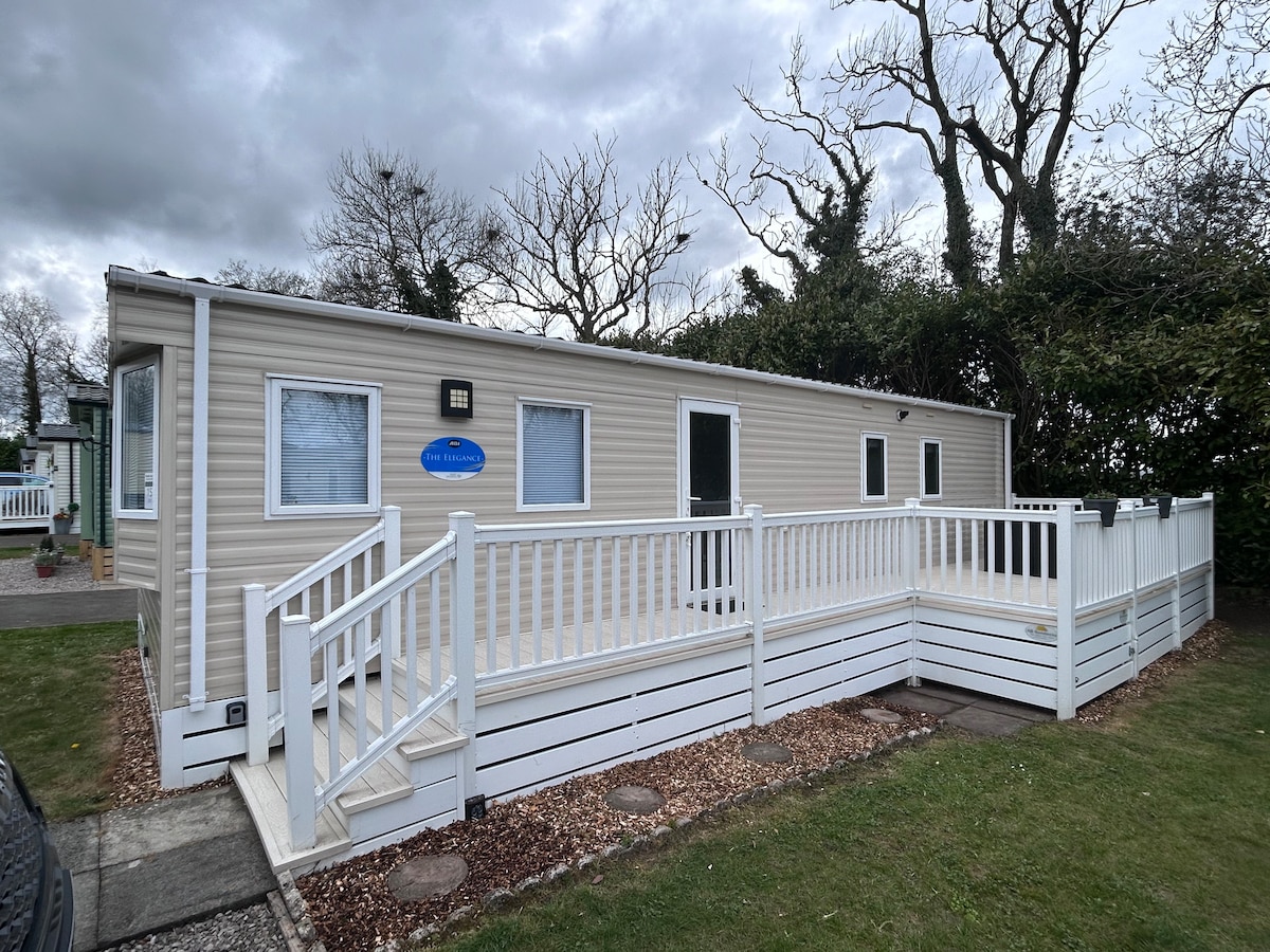 The exterior of a mobile home is displayed, featuring a light-colored facade with multiple windows. A white railing surrounds a spacious deck area, providing access via steps. Surrounding trees and shrubs offer a natural backdrop, while a grassy area is visible in front.