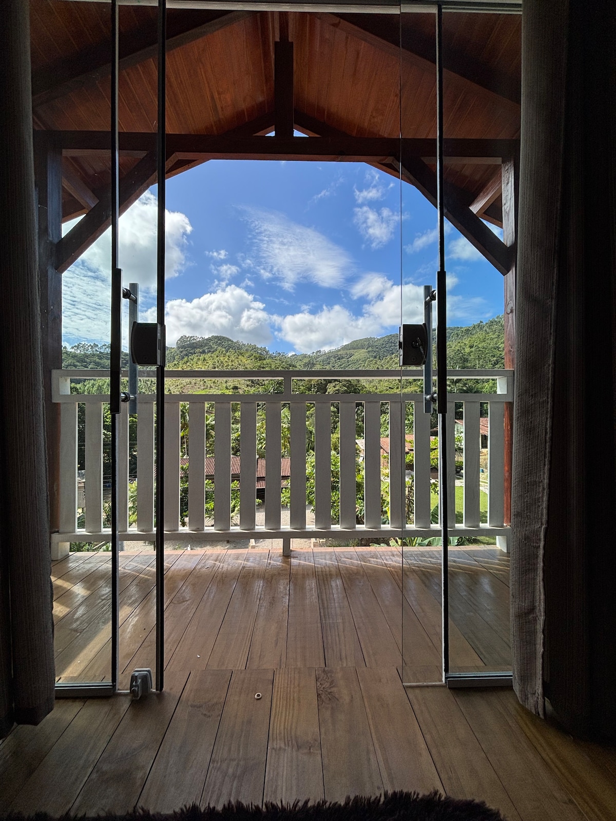 Double glass doors open to reveal a balcony view, framed by wooden beams. The sky is bright with scattered clouds, and lush greenery is visible in the background. The wooden floor adds warmth to the outdoor space, creating a connection with nature.