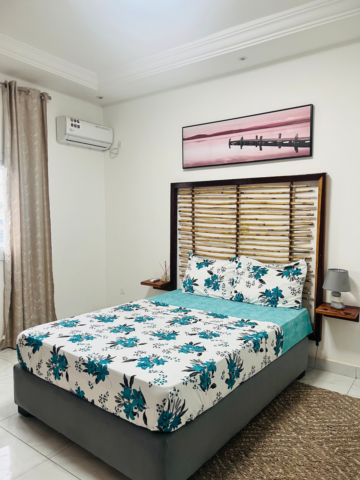 A bedroom features a spacious bed adorned with floral-patterned bedding in shades of blue. Natural wood elements form a decorative headboard, while wall-mounted bedside tables hold lamps. Light enters through a window, complemented by sheer curtains, and a soft area rug enhances the room's comfort.