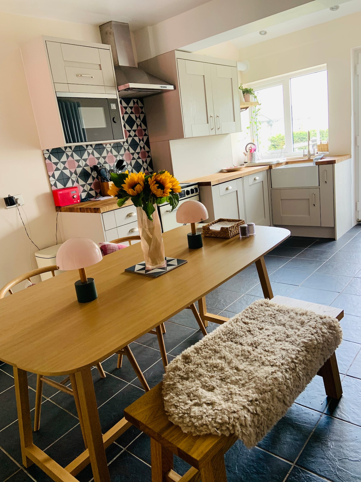 The kitchen features modern cabinetry with a colorful tiled backsplash. A wooden dining table is set with a vase of sunflowers and stylish lamps. Soft textures are present with a woolen throw on the bench. Large windows allow natural light to fill the space.