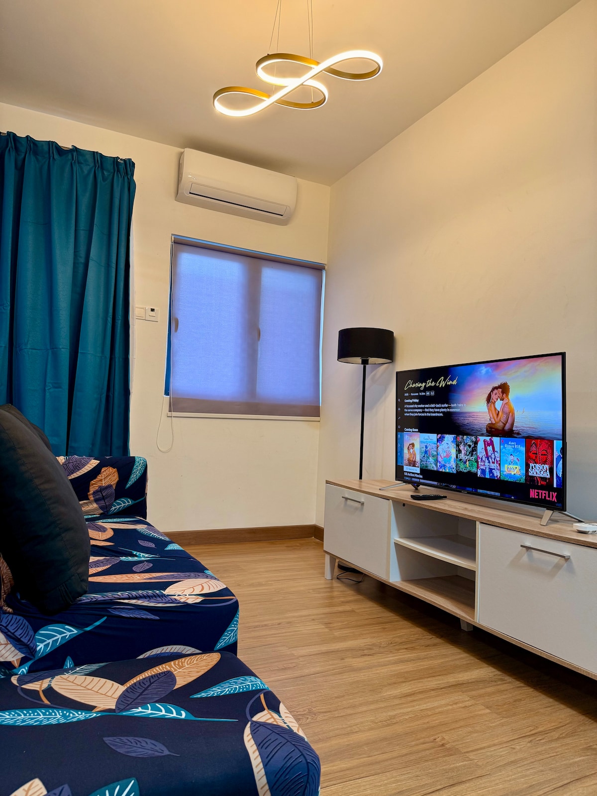 A living area is presented with a vibrant blue sofa adorned with leaf patterns. A modern light fixture hangs above, complemented by a sleek television displaying streaming options. A floor lamp and window are also visible, adding to the functionality of the space.