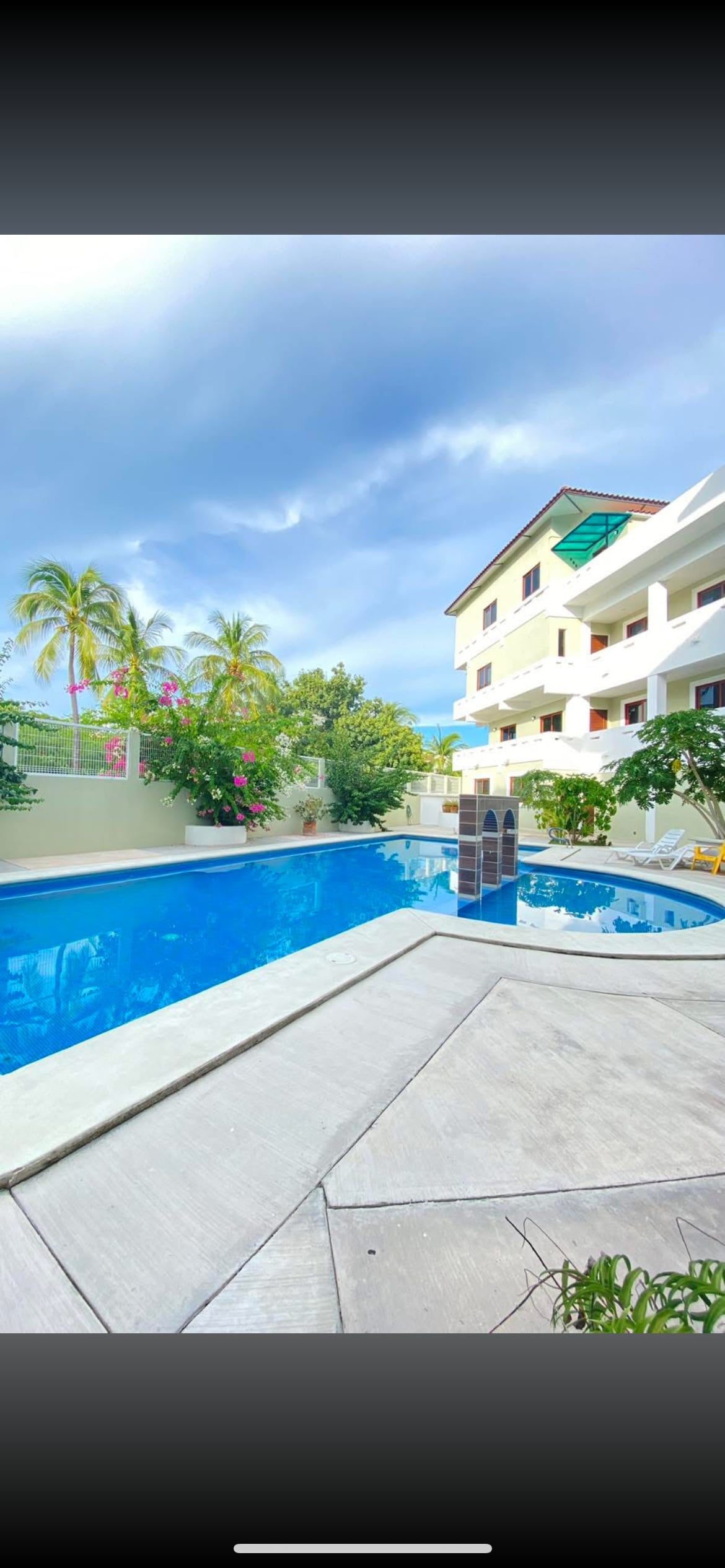 A swimming pool is framed by tropical plants, with lounge chairs positioned nearby. A light-colored building can be seen in the background, featuring multiple balconies adorned with green awnings. The sky showcases varying shades of blue and gray, complementing the serene setting.
