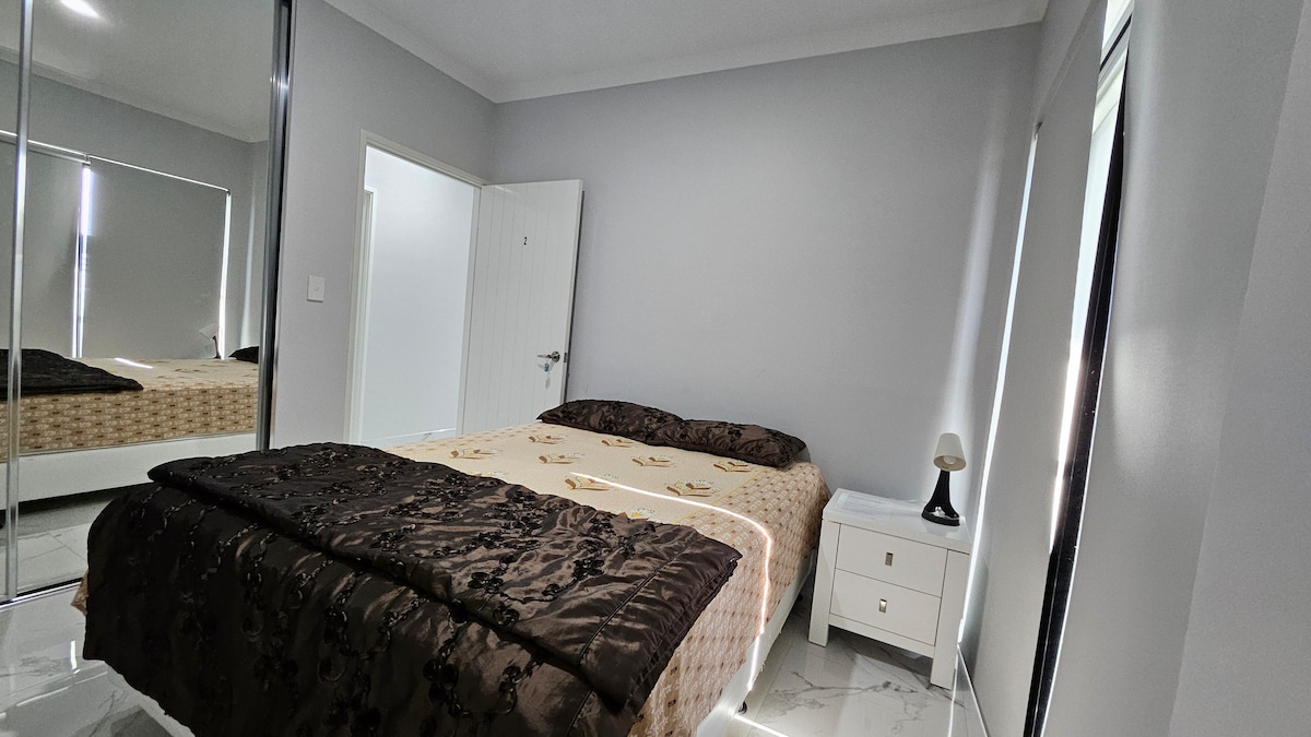 A comfortable bedroom features a spacious bed with an elegantly patterned blanket. Soft lighting is provided by a bedside lamp on a nightstand. A large mirrored wardrobe reflects the room's neutral walls, while natural light enters through partially covered windows.
