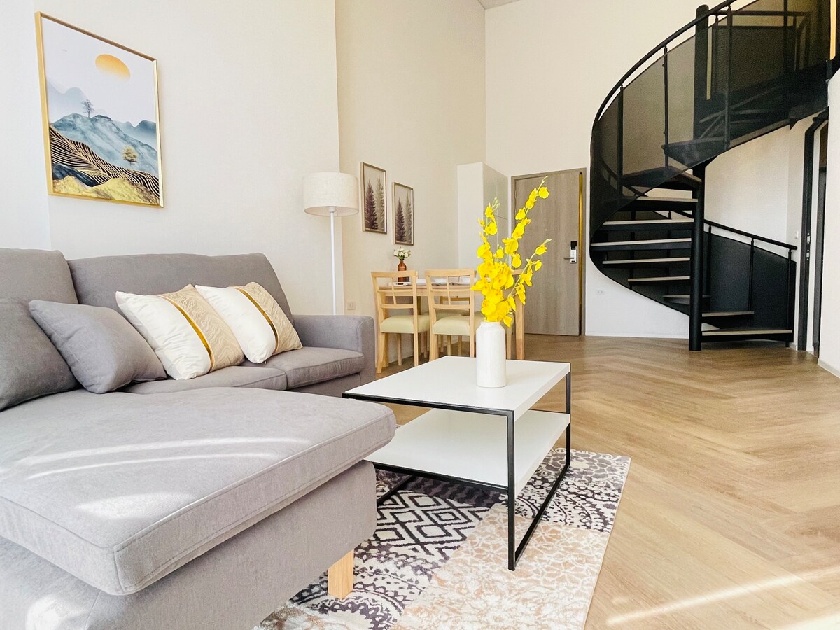 A modern living area features a cozy gray sectional sofa adorned with decorative pillows. A white coffee table sits atop a patterned area rug, while a vase of yellow flowers adds a pop of color. Natural light fills the space, highlighting the spiral staircase in the background.