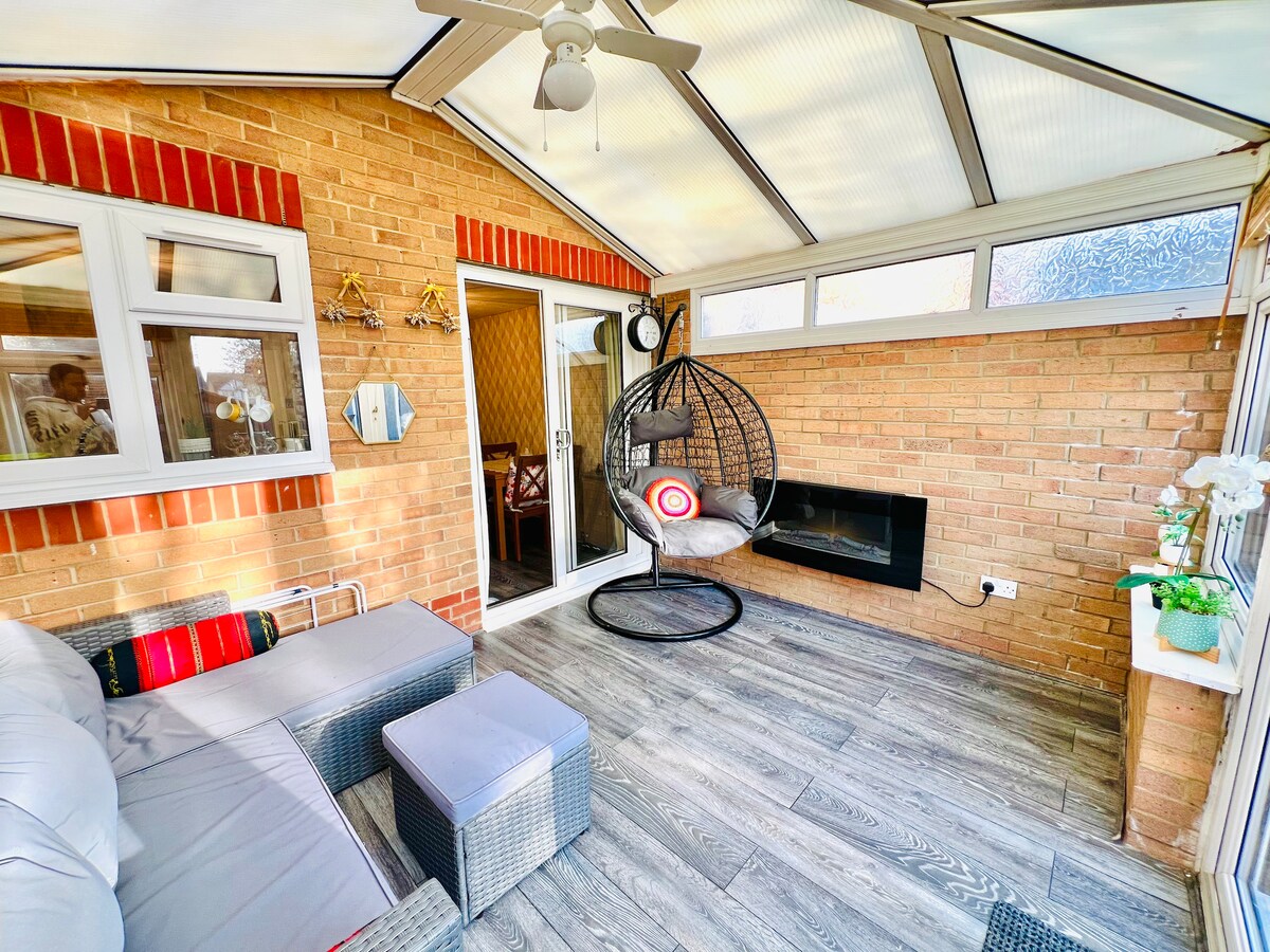 A bright conservatory is showcased with large windows allowing ample natural light. A hanging chair with colorful cushions is positioned near a decorative wall, while a comfortable seating area features a cushioned sofa and a small table. Wooden flooring adds warmth to the space.