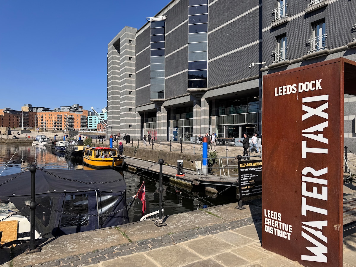 A vibrant canal scene at Leeds Dock showcasing the water taxi station. Yellow water taxis are docked nearby, and several people are seen enjoying the sunny day along the waterfront. The modern buildings of the Leeds Creative District provide a contemporary backdrop.