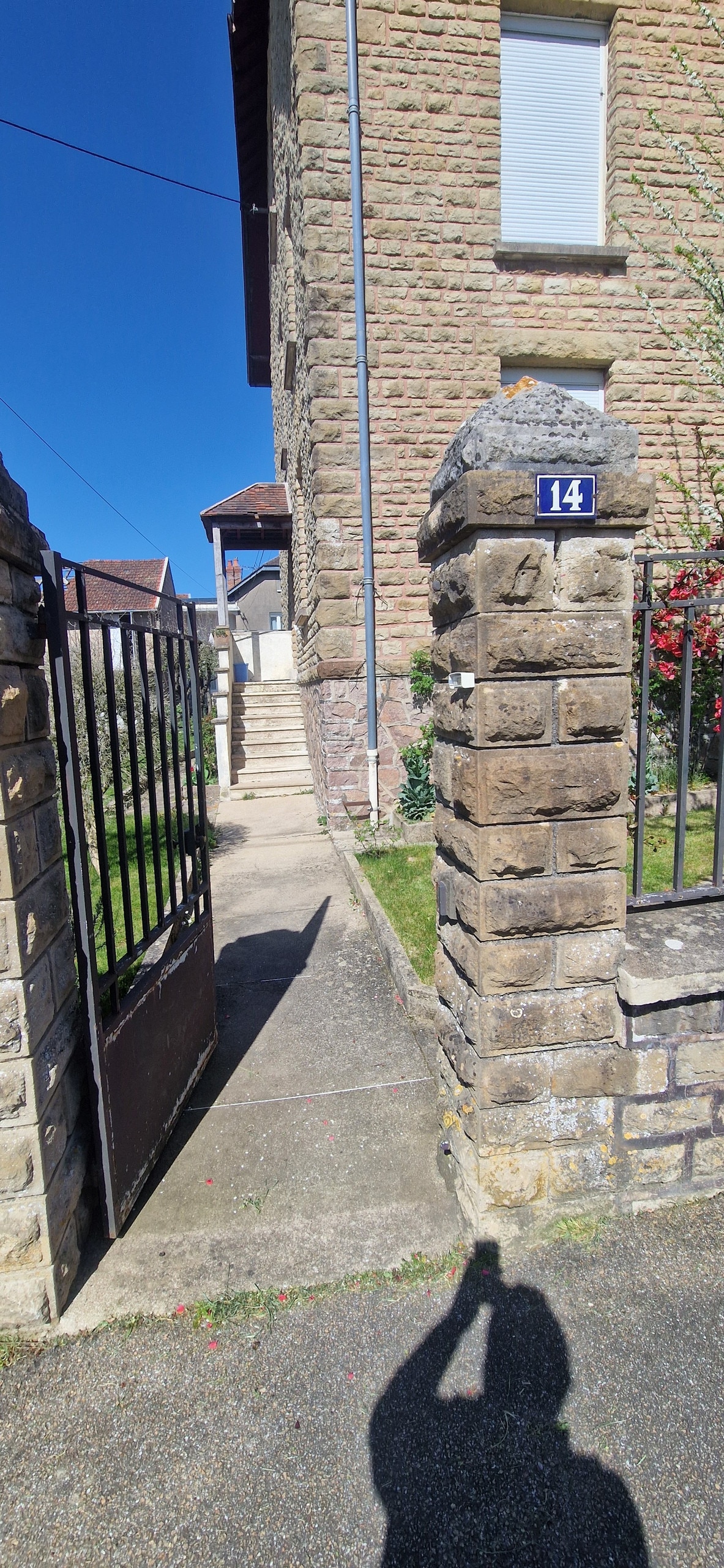A view of a stone entrance gate marked with the number 14 leads to a pathway that extends toward a set of stairs. Lush greenery surrounds the entrance, contributing to a well-maintained exterior. Bright sunlight enhances the clear blue sky above.