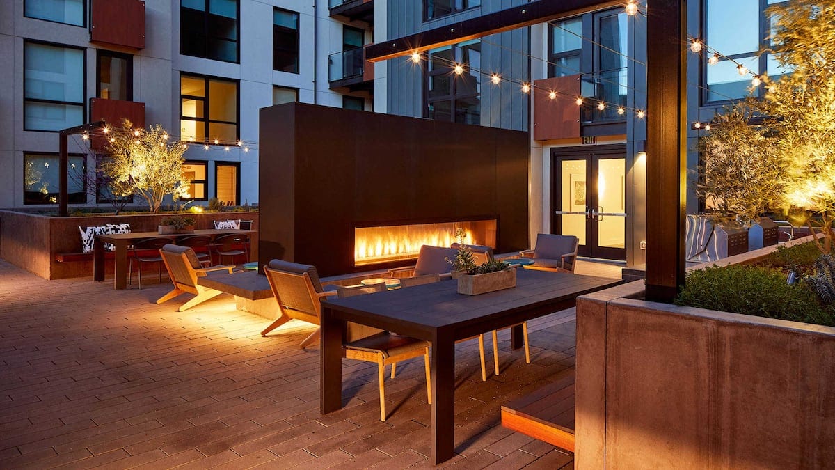 An outdoor communal space features a modern fire pit surrounded by a sleek dark wall. Several wooden lounge chairs and a large dining table are illuminated by soft string lights. Planters with greenery add a touch of nature to the inviting area.