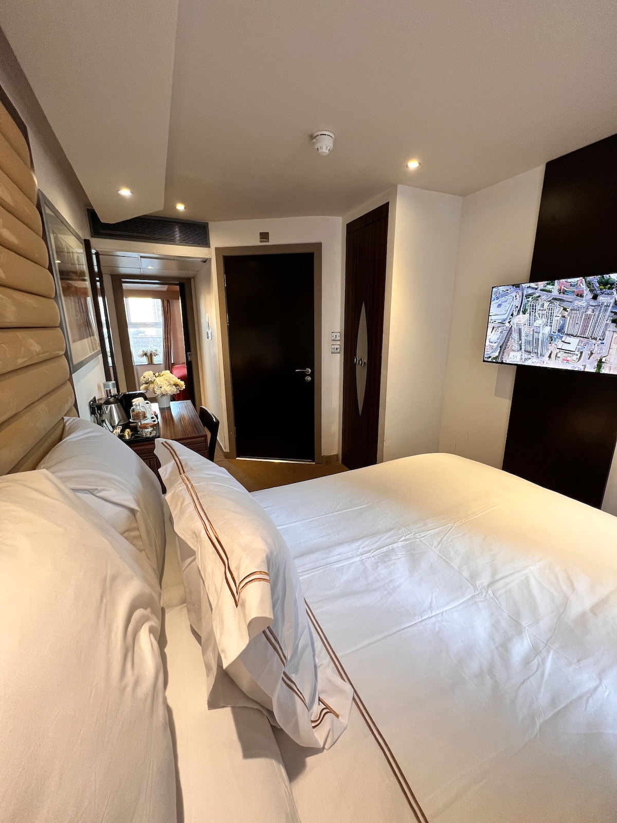 A well-lit bedroom features a spacious bed with crisp white linens. A minimalist design is evident with wooden accents and a mounted TV on the wall. A door leads to a private balcony area, enhancing the room's airy feel.