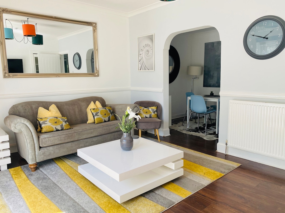 A living area features a comfortable gray sofa adorned with yellow and gray cushions. A sleek white coffee table is centered on a striped rug in yellow and gray tones. A round wall clock and decorative mirror enhance the bright, inviting atmosphere of the space.