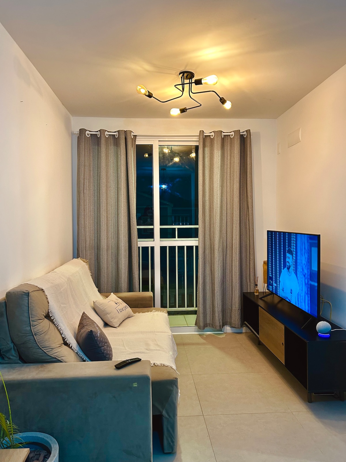 A cozy living area features a light-colored sofa with cushions, positioned across from a television entertainment unit. Natural light enters through a glass door adorned with sheer curtains, creating a comfortable atmosphere. The ceiling light provides warm illumination to the space.