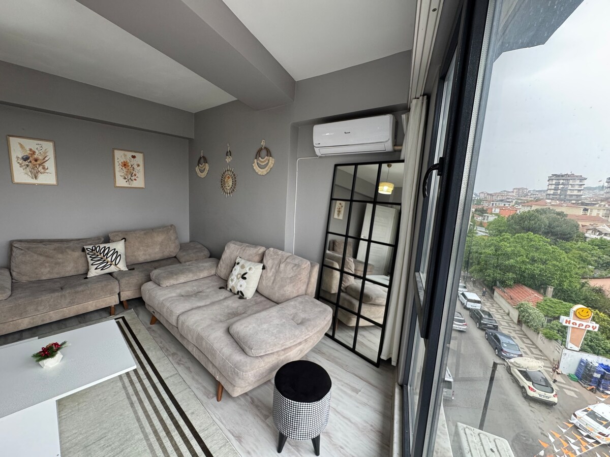 A spacious living area is furnished with a light gray sectional sofa and a black ottoman. A decorative mirror enhances the room's brightness, while wall art adds subtle character. Large windows provide a view of the outside, allowing natural light to fill the space.