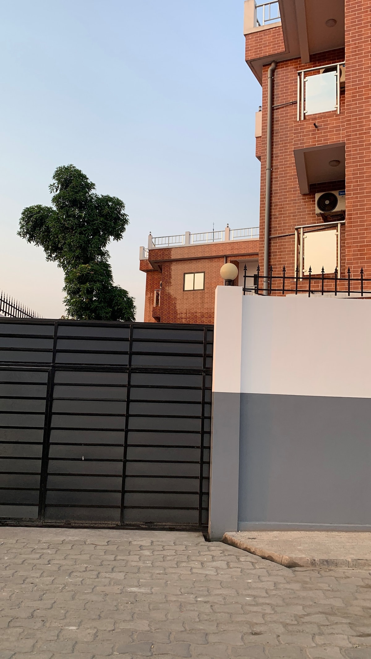 A modern building is viewed from the entrance gate, showcasing a mix of brick and concrete materials. The gate is partially closed, revealing a glimpse of a well-maintained tree nearby. A few balconies can be seen on the upper floors, contributing to the overall façade.