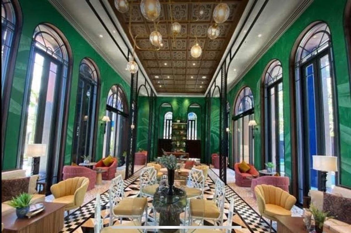 An elegant lobby is characterized by tall, arched windows allowing natural light to illuminate the space. Colorful seating arrangements in shades of yellow and pink complement the vibrant green walls, while a patterned black and white floor adds visual interest. Decorative plants enhance the welcoming environment.