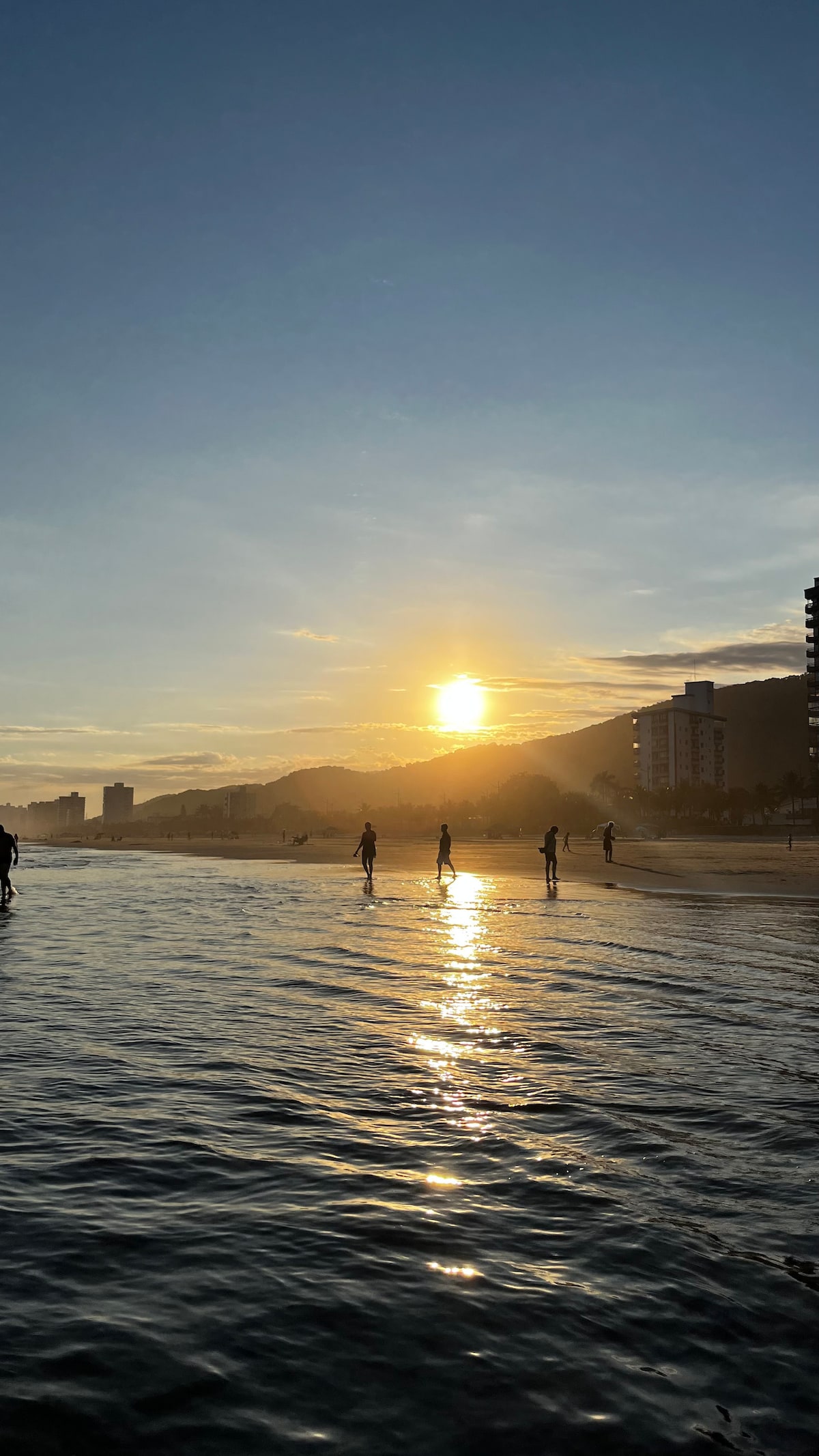 A serene beach scene features a sunset casting soft golden light over gentle waves. Silhouettes of people can be seen strolling along the shore, with distant buildings lining the beach against a backdrop of hills. Clouds diffuse the sunlight, creating a tranquil atmosphere.