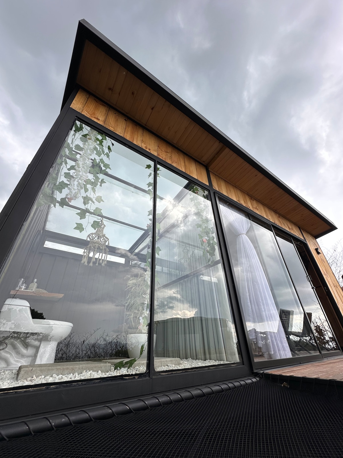 The exterior of a modern building is presented, featuring large glass windows that allow for ample natural light. A wooden overhang complements the architecture, while plants are visible through the glass, creating a connection with nature. A deck area can be seen in the foreground.