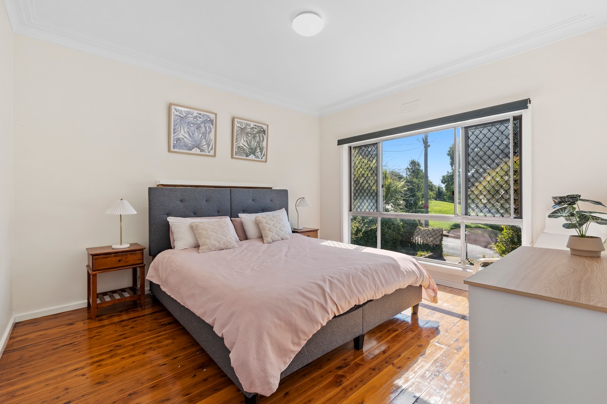 A spacious bedroom features a large bed with a soft pink blanket and cushy pillows. Natural light floods the room through a window adorned with a screen, offering a view of the peaceful exterior. Two artworks hang above the bed, complementing the wooden floor.