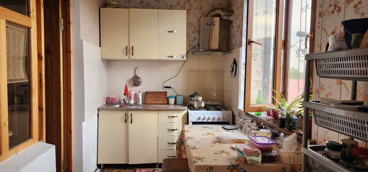 The kitchen features light-colored cabinetry with a tiled countertop, complemented by a gas stove and a sink. Natural light enters through two windows adorned with floral motifs. A dining table is set with a floral tablecloth, surrounded by organized kitchenware and containers.
