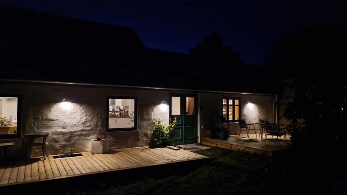The exterior of the house is softly illuminated at night, showcasing a wooden deck with seating options. Two windows allow glimpses of the warmly lit interior, while a green door adds a touch of color against the neutral walls.