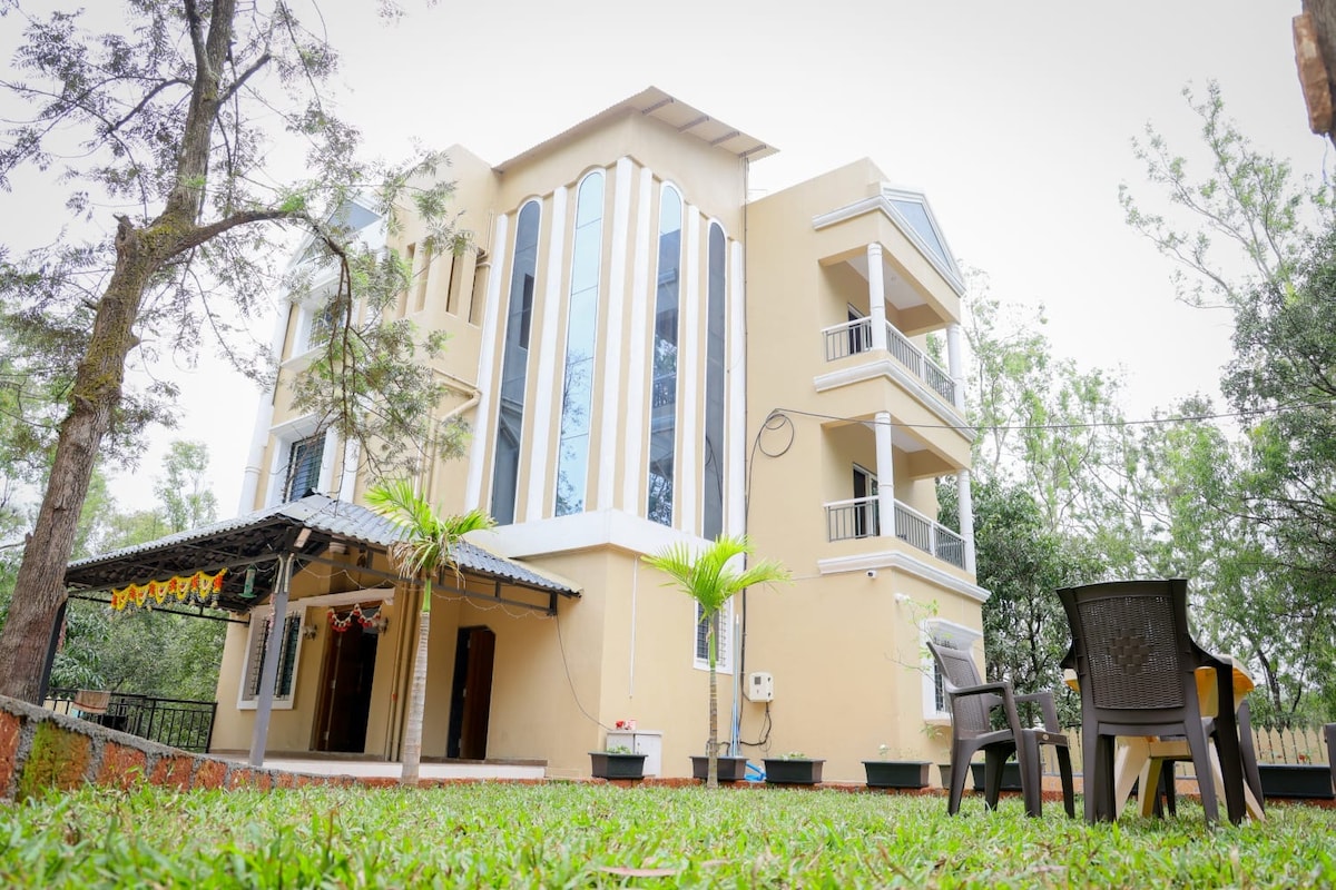 The exterior of the villa features a modern design with large windows and a cream-colored facade. A spacious lawn surrounds the property, and a shaded seating area with chairs is visible. Lush greenery adds to the peaceful ambiance of the location.