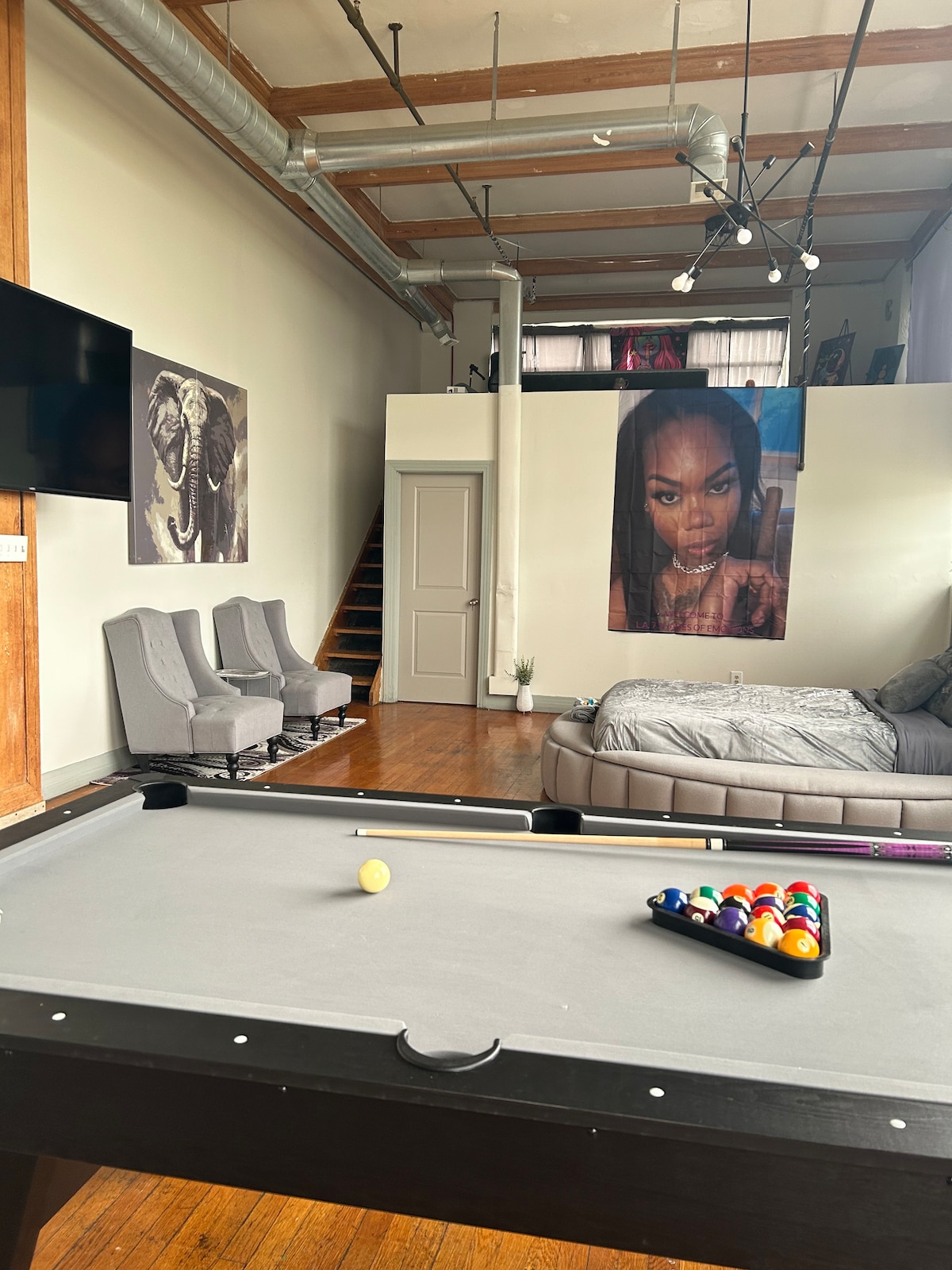 A spacious open area is highlighted by a pool table in the foreground, with a rack of colored balls ready for play. In the background, a comfortable seating arrangement includes two chairs. A modern bed, situated nearby, complements the artistic decor on the walls.