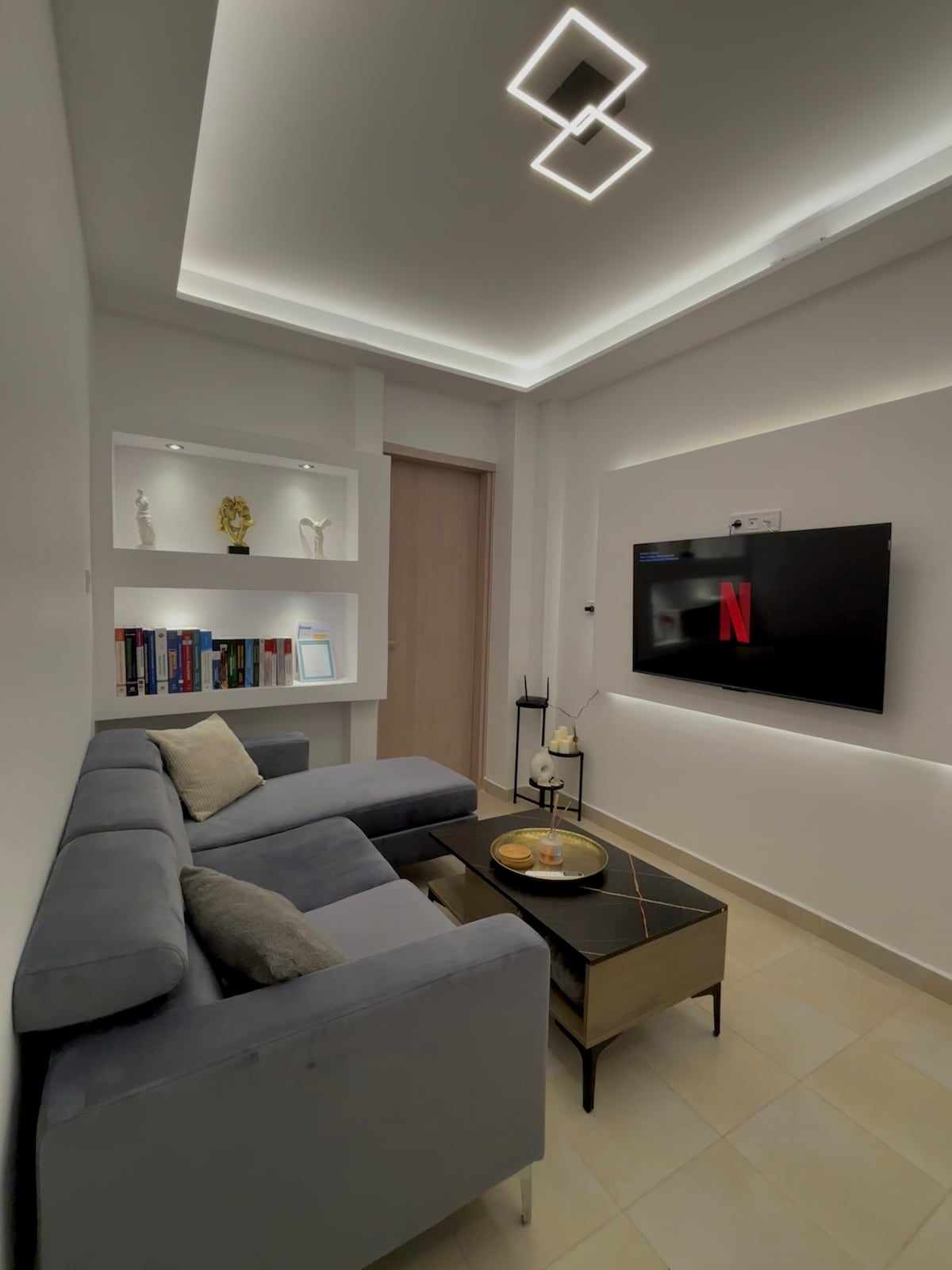 A modern living area is depicted, featuring a gray sofa paired with a sleek coffee table. A wall-mounted television displays the Netflix logo. Shelving units with decorative items and books are integrated into the white walls, illuminated by subtle lighting for visual interest.