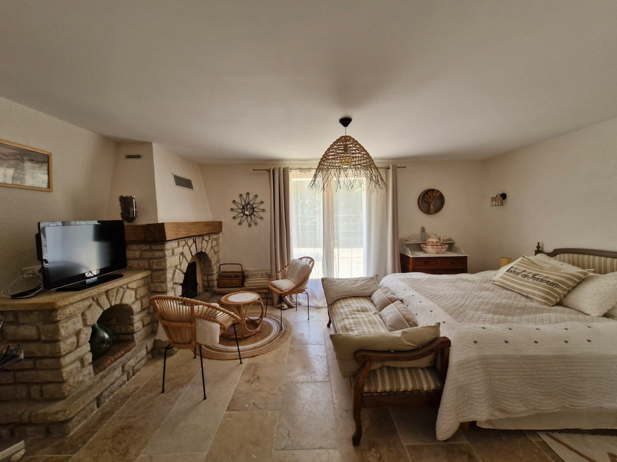 A cozy suite features a large bed and a charming fireplace, accompanied by a comfortable seating area with wicker chairs and a round table. Natural light fills the space through large glass doors, enhancing the warm tones of the stone walls and tiled flooring.